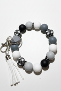 A beaded keychain bracelet featuring matte black, white, and gray round beads, checkered black-and-white beads, a gray heart-shaped bead, and a white faux-leather tassel charm with a silver clasp, displayed on a light off-white background.