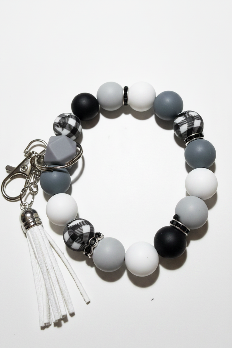 A beaded keychain bracelet featuring matte black, white, and gray round beads, checkered black-and-white beads, a gray heart-shaped bead, and a white faux-leather tassel charm with a silver clasp, displayed on a light off-white background.